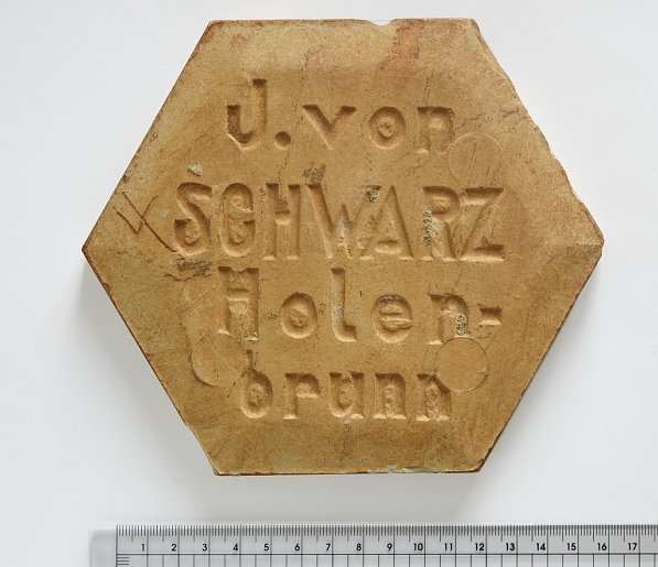 Eine beige Tontafel in der Form eines regelmäßigen Sechseck,
Beschriftung: J. von Schwarz Holenbrunn
am darunter abgebildeten Lineal kann ein Durchmesser von 15 cm abgelesen werden